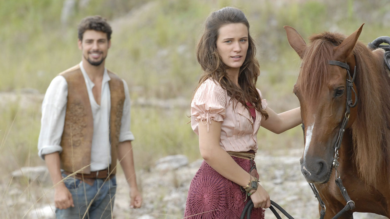 Açucena (Bianca Bin) segura cavalo marrom enquanto Jesuíno (Cauã Reymond) sorri ao fundo em cena externa de Cordel Encantado