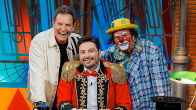 Danilo Gentili sorri de traje vermelho de circo na bancada do The Noite com Luís Ricardo e Palhaço Xuxa em cenário colorido