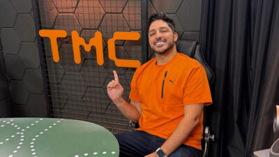 O apresentador Magno Navarro sorrindo e apontando para cima, sentado em estúdio com o logo TMC ao fundo e vestindo camisa laranja