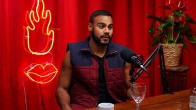 Marcus Brito de colete fala ao microfone no podcast Sem Filtro com cortina vermelha e néon de chama e boca iluminado ao fundo