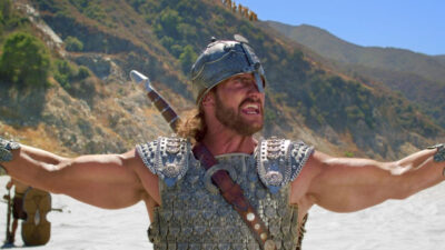 Michael Wayne Foster como Golias grita com braços abertos usando armadura e elmo durante cena do filme Davi & Golias - A Batalha da Fé