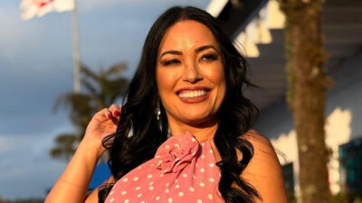 Milene Uehara sorri usando blusa rosa de bolinhas e toca o cabelo em área externa do SBT