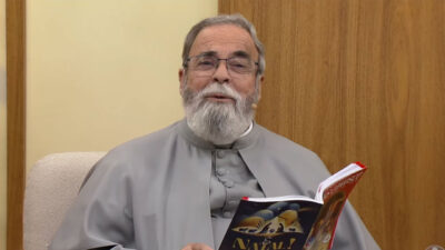 Padre Antonio Maria de batina cinza e barba branca segura livro de Natal sentado em poltrona bege nos estúdios da TV Aparecida
