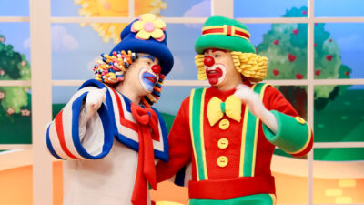 Os palhaços Patati e Patatá, com fantasias coloridas, sorrindo um para o outro durante gravação no cenário do Bom Dia & Cia