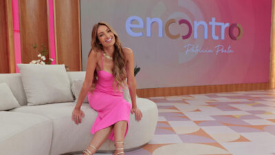 Patrícia Poeta sorri sentada no sofá branco do Encontro usando vestido rosa com logotipo do programa no telão ao fundo do estúdio