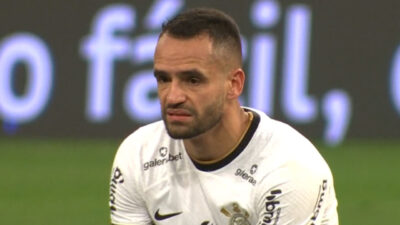 Renato Augusto com expressão séria veste camisa branca do Corinthians durante partida de futebol com painel de LED ao fundo no estádio