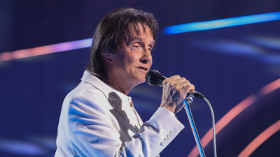 O cantor Roberto Carlos, vestindo um terno branco, canta segurando um microfone durante especial da TV Globo