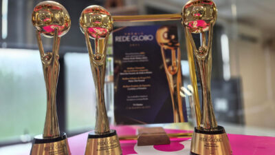 Três troféus dourados do Prêmio Rede Globo em destaque sobre base rosa com placa comemorativa da RPC ao fundo em ambiente interno