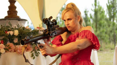 Rebel Wilson como Sam segura uma bazuca com expressão séria e vestido rosa em cena de casamento do filme de ação Duro de Casar