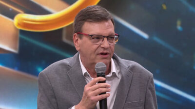 Narrador esportivo Sergio Maurício, de óculos e blazer cinza, fala ao microfone durante gravação de programa em estúdio de TV