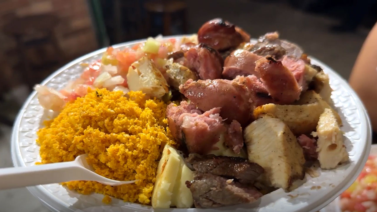 Um prato de plástico branco cheio de comida, contendo farofa amarela, vinagrete (tomate picado com cebola), pedaços de linguiça grelhada ou assada, além de pedaços de carne. Uma colher de plástico branca está na farofa, apontando para fora da imagem