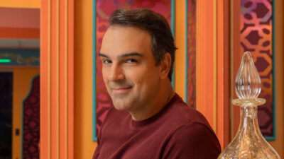 Apresentador Tadeu Schmidt com camiseta vinho sorri discretamente em cenário com paredes coloridas do Big Brother Brasil, da Globo