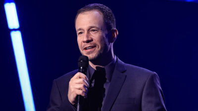 Tiago Leifert de terno escuro fala ao microfone com leve sorriso em palco de programa de TV iluminado por luzes azuis ao fundo