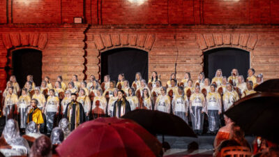 Grande coral veste capas de chuva transparentes durante apresentação da Cantata de Natal em frente a um cenário de tijolos iluminado de vermelho, com público segurando guarda-chuvas