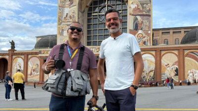 Willian Corrêa e Chico Regueira posam sorrindo com câmera em frente à fachada do Santuário Nacional de Aparecida em reportagem