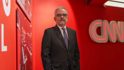 Antonio Lavareda de terno escuro e óculos posa em frente ao logotipo da CNN Brasil em um corredor com paredes vermelhas da emissora