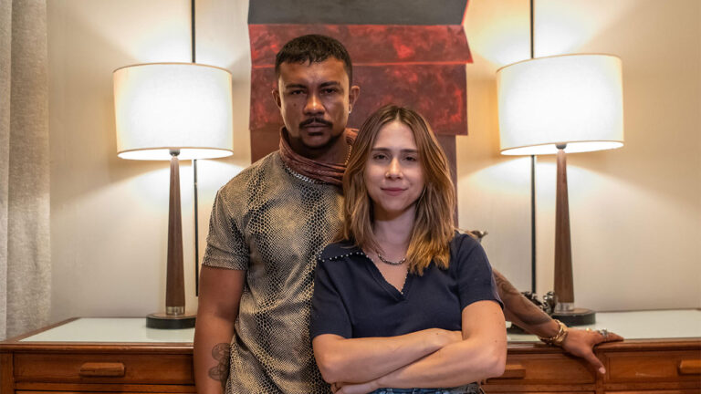 Bagdá (Xamã) e Lucélia (Daphne Bozaski) em ambiente interno da novela Três Graças com ela de braços cruzados e ele atrás em pose séria