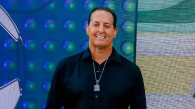 Benjamin Back sorridente com camisa preta e colar de placa no cenário do programa Arena SBT diante de painel tecnológico azul e verde