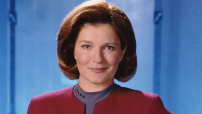 Kate Mulgrew como Capitã Kathryn Janeway com uniforme da Frota Estelar em fundo azul em cena da série Jornada nas Estrelas - Voyager