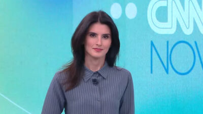 Carol Nogueira apresenta o CNN Novo Dia em estúdio da CNN Brasil com camisa cinza listrada e fundo azul com logo do jornal