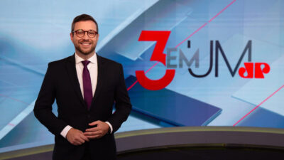 Cassius Zeilmann de óculos e gravata roxa sorri com as mãos unidas à frente do logo do programa 3 em 1 na Jovem Pan