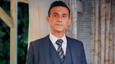 Cem Bender como Celebi de terno azul e gravata listrada com expressao seria em cena da novela turca Chamas do Destino