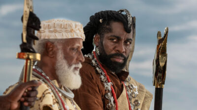 Chinua (Hilton Cobra) e Jendal (Lázaro Ramos) lado a lado com trajes de época e bastões cerimoniais em cena da novela A Nobreza do Amor