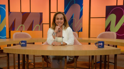 Cissa Guimarães sorri sentada na bancada do Sem Censura usando blazer branco com caneca azul no cenário colorido da TV Brasil