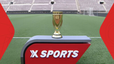 Troféu dourado da Copinha sobre bancada vermelha com logotipo Xsports em campo de futebol vazio com arquibancadas claras ao fundo