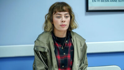 Demet Evgar como Cemre Kayabeyli em Chamas do Destino veste parka verde e camisa xadrez com expressão séria em ambiente azulado