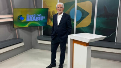 Domingos Meirelles de terno azul e camisa branca posa no cenário do programa Destino Brasil TV nos estúdios da TV Serra Dourada