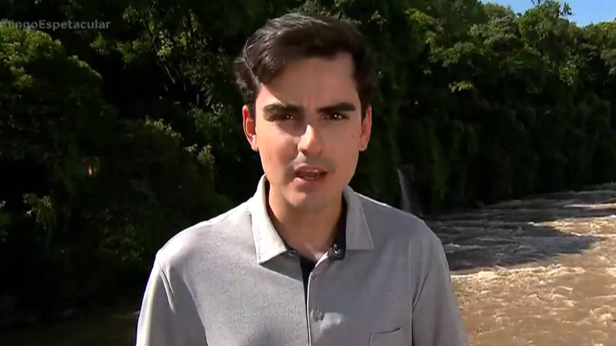 Dudu Camargo fala em área externa com camisa cinza diante de vegetação e curso de água ao fundo durante gravação de reportagem