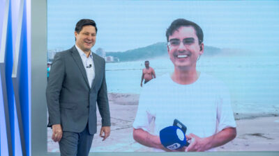 Eleandro Passaia de terno cinza no estúdio do Balanço Geral sorri ao lado de telão com Dudu Camargo em reportagem externa na praia