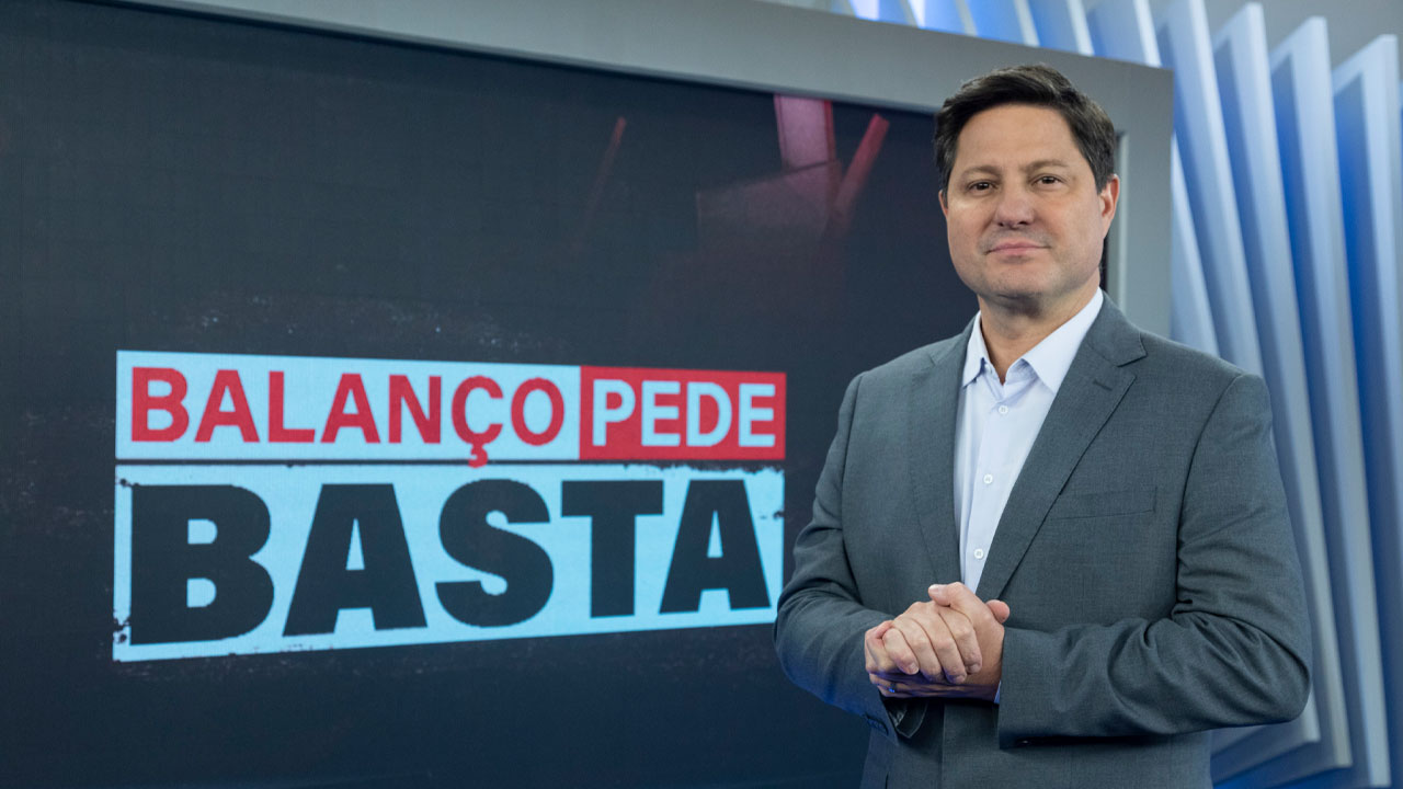Eleandro Passaia de terno cinza e camisa clara posa com as mãos entrelaçadas à frente do telão com o selo Balanço Pede Basta no estúdio