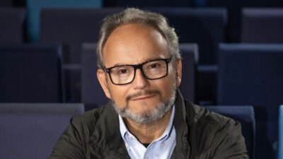 Jornalista Ernesto Paglia com óculos e barba grisalha sentado em poltrona de cinema vazia olhando para a câmera