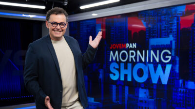 Fernando Rocha sorri de óculos azuis e blazer escuro enquanto gesticula ao lado do logo do Morning Show no estúdio da Jovem Pan