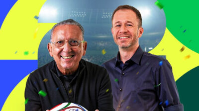 Galvão Bueno e Tiago Leifert posam sorrindo com bola de futebol em estúdio com fundo gráfico nas cores verde amarelo e azul