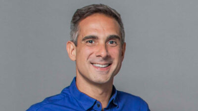 Gustavo Villani sorri para a foto usando camisa social azul em fundo cinza neutro durante ensaio oficial de narradores da Globo