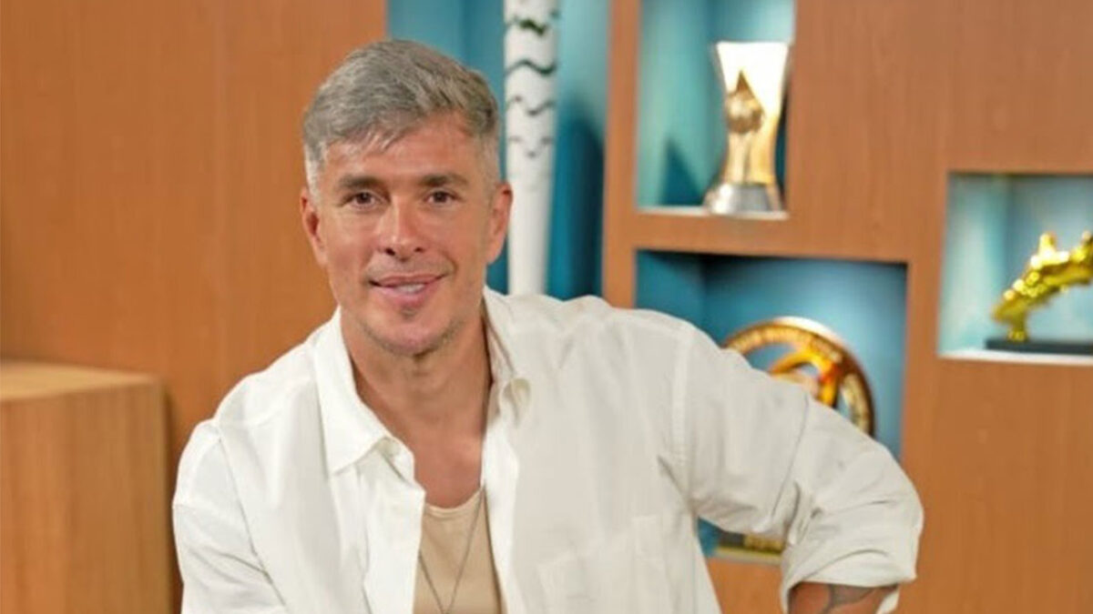 Ivan Moré sorri com cabelos grisalhos curtos usando camisa branca aberta sobre camiseta bege em estúdio com troféus de futebol ao fundo