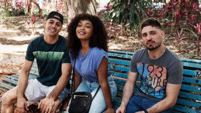 Jean Amorim com boné preto, Larissa Nunes de camiseta azul e Daniel Rocha com blusa cinza sentados em banco de praça no filme Nossa Vizinhança