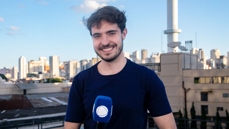 João Liberato sorri segurando microfone da Record em área externa com prédios ao fundo durante gravação de reportagem