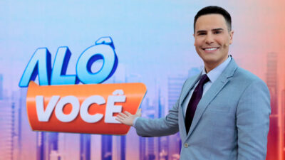 Luiz Bacci sorri de terno cinza e gravata roxa enquanto aponta para o logotipo do programa Alô Você em estúdio com fundo de prédios estilizados