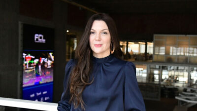 Mafê Bertone com blusa de cetim azul marinho e cabelos longos escuros posa nos corredores da TV Gazeta em frente ao painel da FCL