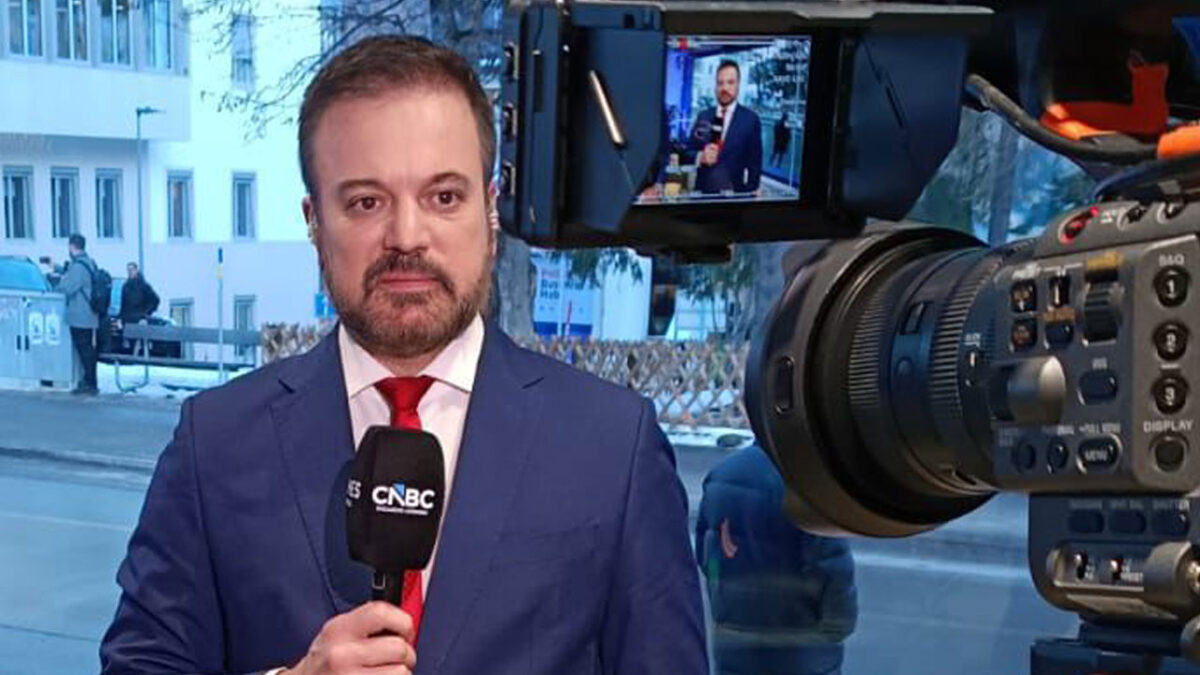 Marcelo Torres com terno azul e gravata vermelha segura microfone da CNBC enquanto é filmado por câmera em área interna com prédio ao fundo na janela de vidro