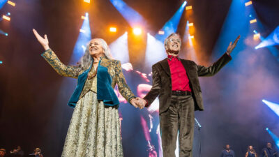 Maria Bethânia e Caetano Veloso sorriem de mãos dadas com braços erguidos sob luzes de palco durante show da turnê Caetano e Bethânia