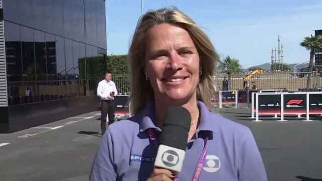Mariana Becker sorri segurando microfone da TV Globo com logotipo da emissora durante cobertura de Fórmula 1 em um autódromo