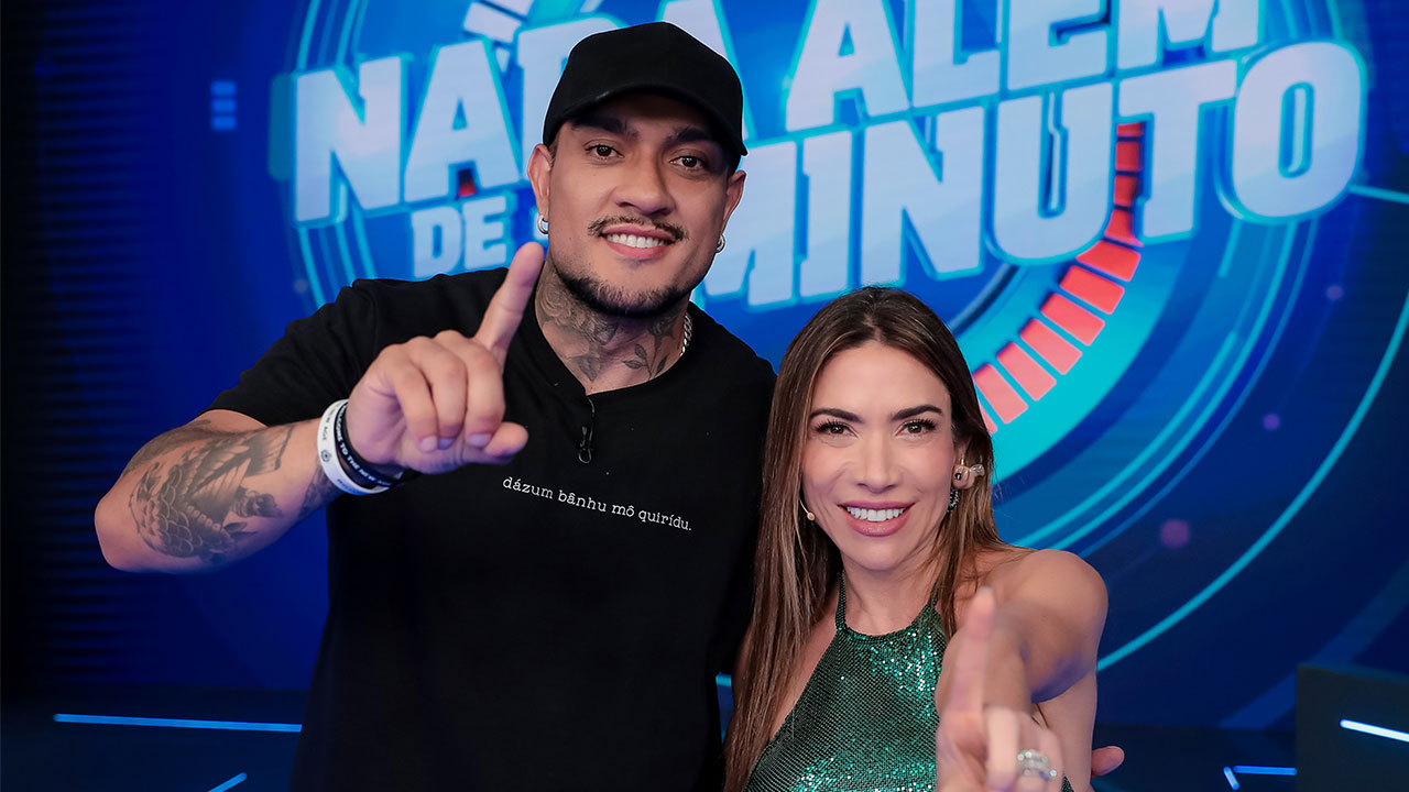 MC Binn e Patricia Abravanel posam no palco do Programa Silvio Santos com ambos levantando o dedo indicador diante do telão com logo do Nada Além de 1 Minuto