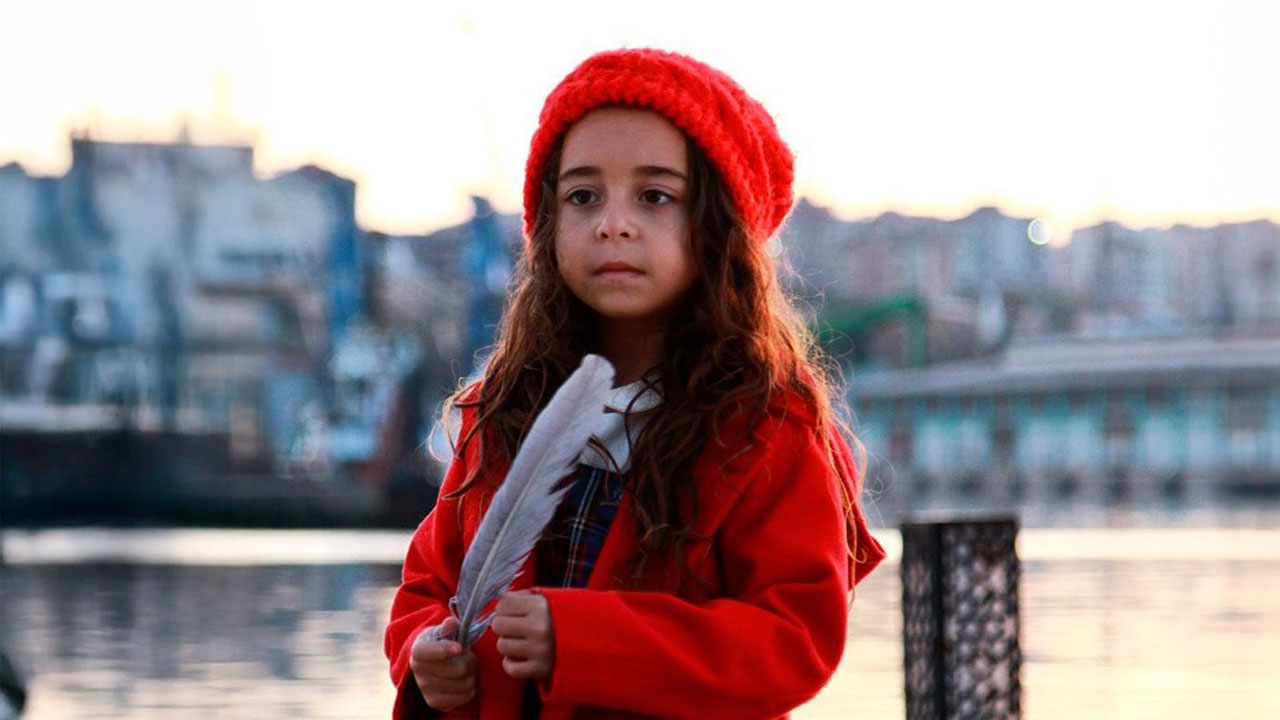 Beren Gokyildiz como Melek segura uma pena branca vestindo casaco e gorro vermelhos em cena da novela turca Mãe com mar ao fundo