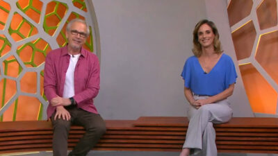 Nélson Araújo e Cristina Vieira sorriem sentados no banco de madeira do cenário do Globo Rural com painéis hexagonais ao fundo na TV Globo