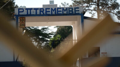 Entrada da Penitenciária de Tremembé II com letreiro azul em arco branco vista através de grade desfocada com placas de sinalização e árvores ao fundo